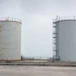 Two steel storage tanks at a coastal site, one with weathered paint and rust along the weld seams, the other recoated in light grey
